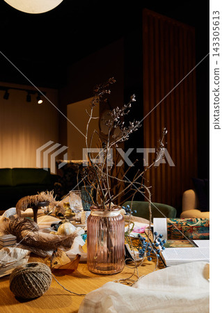 Dried twig arrangement in ribbed glass vase on cluttered wooden table with craft supplies, warm indoor lighting and moody vertical wood panel background Dried twig arrangement in ribbed glass vase on cluttered wooden table with craft supplies, warm indoor lighting and moody vertical wood panel background 143305613