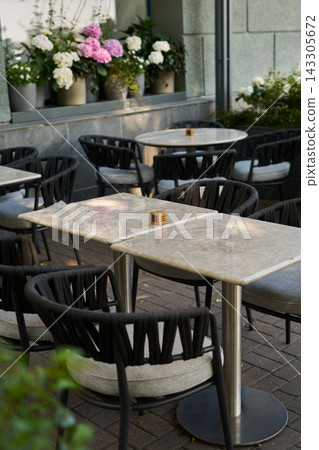 Outdoor cafe tables with woven rope chairs, potted hydrangeas on windowsill, marble tabletops on brick pavement, cozy urban terrace for restaurant or hospitality use Outdoor cafe tables with woven rope chairs, potted hydrangeas on windowsill, marble tabletops on brick pavement, cozy urban terrace for restaurant or hospitality use 143305672