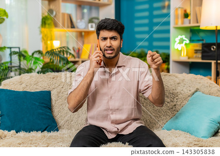 Indian man on home sofa arguing on smartphone with stressed talk using hand gesture during live call Indian man on home sofa arguing on smartphone with stressed talk using hand gesture during live call 143305838