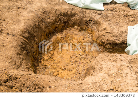 A freshly dug hole in the ground shows prepared soil for planting purposes, surrounded by loose earth. A freshly dug hole in the ground shows prepared soil for planting purposes, surrounded by loose earth. 143309378