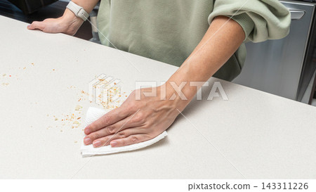 Woman wiping crumbs from kitchen table with hand, homemade baking process, food preparation routine, cozy home cooking lifestyle. Woman wiping crumbs from kitchen table with hand, homemade baking process, food preparation routine, cozy home cooking lifestyle. 143311226