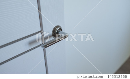 Sleek chrome handle mounted on white interior door, shown in soft daylight with minimal lines and shallow depth of field. Quiet home or office entrance detail with clean modern style. 143312366