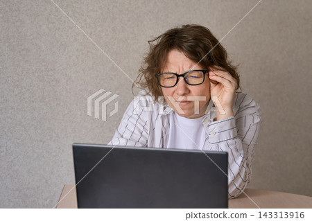 Woman Adjusting Glasses While Leaning Close To Laptop At Table In Plain Indoor Room With Beige Wall Woman Adjusting Glasses While Leaning Close To Laptop At Table In Plain Indoor Room With Beige Wall 143313916