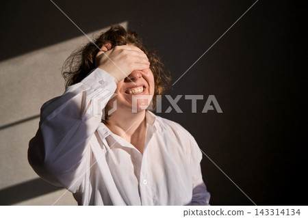 Adult Woman Covering Eyes And Grimacing From Pain In White Shirt Under Strong Side Light Against Adult Woman Covering Eyes And Grimacing From Pain In White Shirt Under Strong Side Light Against 143314134