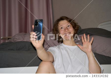 Woman Waves Spread Fingers Beside Vertically Held Phone While Smiling Toward Screen, With Bent Knee Woman Waves Spread Fingers Beside Vertically Held Phone While Smiling Toward Screen, With Bent Knee 143314192