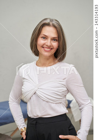 Smiling Woman Standing In White Layered Top And Black Trousers Inside Calm Treatment Room Smiling Woman Standing In White Layered Top And Black Trousers Inside Calm Treatment Room 143314193
