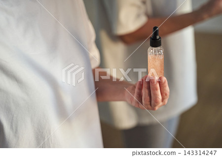 Woman Presenting Glass Dropper Bottle Of Glittering Serum In Soft Light Near Mirrored Companion Woman Presenting Glass Dropper Bottle Of Glittering Serum In Soft Light Near Mirrored Companion 143314247