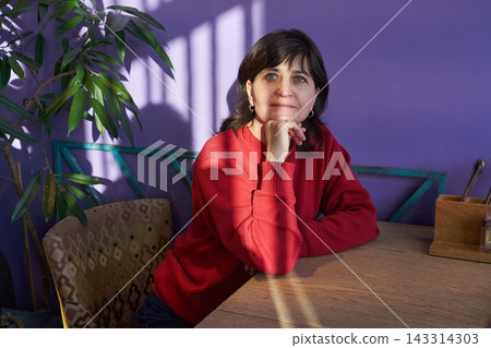 Adult Woman Resting Chin On Hand Seated At Wooden Table In Sunlit Interior With Purple Wall Adult Woman Resting Chin On Hand Seated At Wooden Table In Sunlit Interior With Purple Wall 143314303
