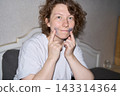 Woman Touching Blue Under Eye Patches With Both Index Fingers While Smiling At Camera In Bedroom 143314364