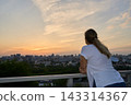 Young Woman Leaning On Balcony Railing Watching Distant City Skyline Under Warm Sunset Clouds 143314367