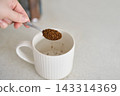 Spoon Releasing Instant Coffee Granules Into White Mug On Kitchen Counter With Hand Visible And Jar 143314369