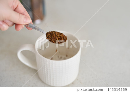 Spoon Releasing Instant Coffee Granules Into White Mug On Kitchen Counter With Hand Visible And Jar Spoon Releasing Instant Coffee Granules Into White Mug On Kitchen Counter With Hand Visible And Jar 143314369