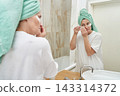 Woman Adjusting Under Eye Patches In Front Of Bathroom Mirror With Towel Wrapped Hair Standing 143314372