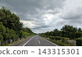 A highway surrounded by greenery stretches straight ahead beneath thick clouds. 143315301