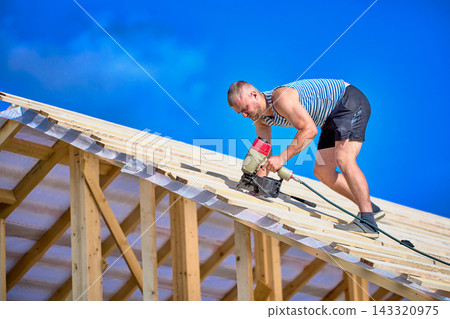 Securing timber battens over roofing underlay, worker uses nail gun while assembling wooden roof structure during construction. Securing timber battens over roofing underlay, worker uses nail gun while assembling wooden roof structure during construction. 143320975