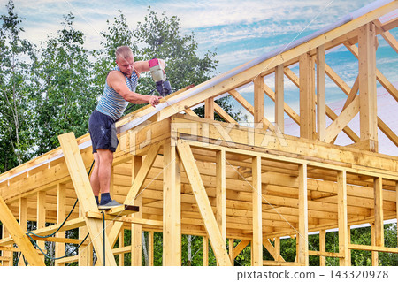 Carpenter fastens roof laths over weather barrier using compressed air nailer while standing on makeshift work platform. Carpenter fastens roof laths over weather barrier using compressed air nailer while standing on makeshift work platform. 143320978