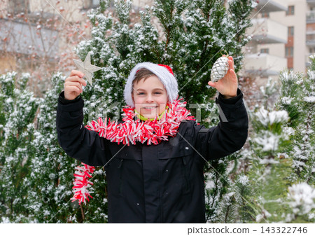 Child dressed as Santa with white beard and red tinsel garland standing outdoors Child dressed as Santa with white beard and red tinsel garland standing outdoors 143322746