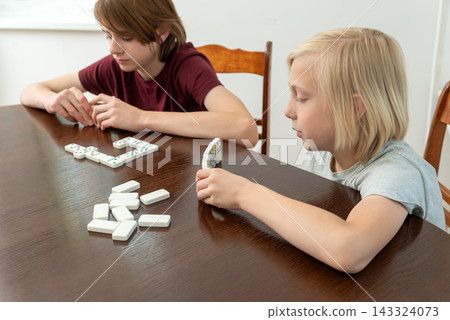 Two young children are thoughtfully engaging in a game of dominoes at a wooden table, concentrating on placing the tiles and enjoying a quiet indoor activity Two young children are thoughtfully engaging in a game of dominoes at a wooden table, concentrating on placing the tiles and enjoying a quiet indoor activity 143324073