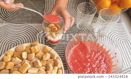 Woman hands holding spoon adding strawberry puree syrup over savoiardi biscuits inside glass for making eggless tiramisu dessert process top view Woman hands holding spoon adding strawberry puree syrup over savoiardi biscuits inside glass for making eggless tiramisu dessert process top view 143324816