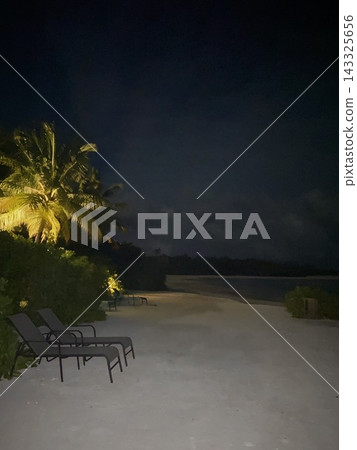 Empty lounge chairs and illuminated palm trees line a tropical beach at night. Empty lounge chairs and illuminated palm trees line a tropical beach at night. 143325656