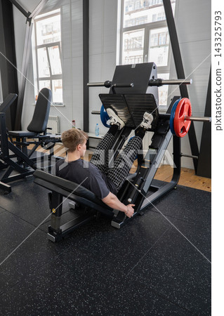 Young man training legs on angled press machine in bright modern gym, pushing weighted sled with white athletic shoes while seated against black padding near large daylight windows Young man training legs on angled press machine in bright modern gym, pushing weighted sled with white athletic shoes while seated against black padding near large daylight windows 143325793