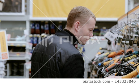 Customer in a hardware store carefully selecting an electric drill while wearing a black jacket. A man in a hardware store choosing a tool. Customer in a hardware store carefully selecting an electric drill while wearing a black jacket. A man in a hardware store choosing a tool. 143325840
