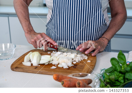 Senior woman methodically dicing white onions on a wooden board in a sunlit kitchen 143327353
