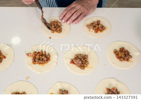 Woman portioning spiced meat filling onto empanada dough rounds with focused efficiency Woman portioning spiced meat filling onto empanada dough rounds with focused efficiency 143327391