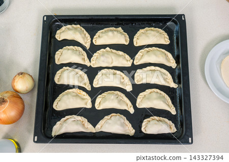 Fifteen unbaked empanadas arranged in neat rows on a dark baking tray ready for the oven Fifteen unbaked empanadas arranged in neat rows on a dark baking tray ready for the oven 143327394