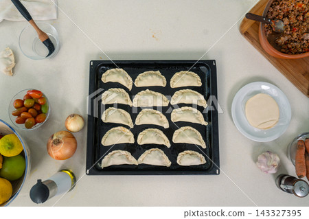 Unbaked empanadas arranged in orderly rows on a dark baking sheet surrounded by fresh ingredients Unbaked empanadas arranged in orderly rows on a dark baking sheet surrounded by fresh ingredients 143327395