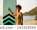 Black woman holding paddleboard on beach looking out at ocean 143328560