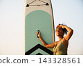 Black woman holding paddleboard on beach looking out at ocean 143328561