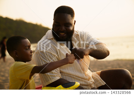 Black father sitting with son on beach talking during sunset Black father sitting with son on beach talking during sunset 143328566