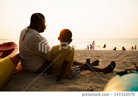 Black father sitting with son on beach talking during sunset 143328570