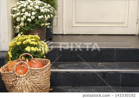 Autumn entrance decorated with chrysanthemums and pumpkins in a woven basket, creating a cozy seasonal scene with rustic charm and Halloween atmosphere. Autumn entrance decorated with chrysanthemums and pumpkins in a woven basket, creating a cozy seasonal scene with rustic charm and Halloween atmosphere. 143330192