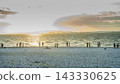 A photographic image of people gazing at the sea on a beach at sunset, with the water's surface shimmering, the white pebble beach, and the setting sun. 143330625