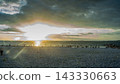A photograph of a white pebble beach at sunset, with the setting sun shining through a break in the clouds and people standing by. 143330663