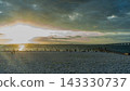 A photographic image of a coastline at dusk, with the setting sun shining through the clouds, showcasing the white pebble beach and the connecting bridge. 143330737