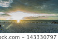 A photograph of a white pebble beach at sunset, with the setting sun shining through a break in the clouds and people standing by. 143330797
