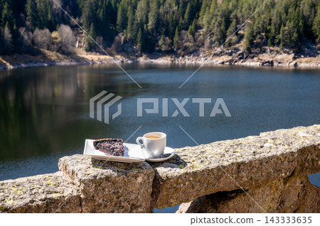 Fresh pastry and espresso cup resting on stone railing with distant pine reflections on calm lake, quiet lakeside breakfast moment in mountain setting 143333635
