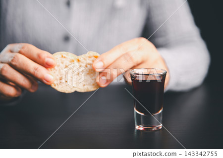 Person breaking bread beside a small glass of dark red communion beverage on a black table, representing Christian worship, sacred remembrance, faith, prayer, fellowship, sacrifice, and reflection. Person breaking bread beside a small glass of dark red communion beverage on a black table, representing Christian worship, sacred remembrance, faith, prayer, fellowship, sacrifice, and reflection. 143342755