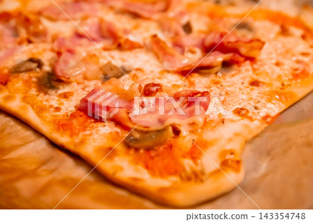 Baked cheese pizza topped with ham and vegetables on a wooden board. Close-up view of savory pizza for casual dining. Italian cuisine, comfort food and meal sharing concept. Baked cheese pizza topped with ham and vegetables on a wooden board. Close-up view of savory pizza for casual dining. Italian cuisine, comfort food and meal sharing concept. 143354748