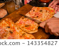 Hands slicing pizza with a kitchen knife on wooden board. Shared pizza meal and food preparation. Italian cuisine, casual dining and home cooking. Group dining and meal sharing. 143354827