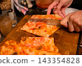 Hands slicing pizza with a kitchen knife on wooden board. Shared pizza meal and food preparation. Italian cuisine, casual dining and home cooking. Group dining and meal sharing. 143354828