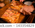 Hands slicing pizza with a kitchen knife on wooden board. Shared pizza meal and food preparation. Italian cuisine, casual dining and home cooking. Group dining and meal sharing. 143354829