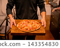 Person carrying a baked pizza on a wooden serving board in a kitchen. Italian cuisine and casual dining. Food service and restaurant hospitality. Pizza dinner for sharing and entertaining. 143354830