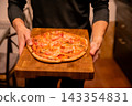 Man carrying a baked pizza on a wooden serving board in a kitchen. Italian cuisine and casual dining. Food service and restaurant hospitality. Pizza dinner for sharing and entertaining. 143354831