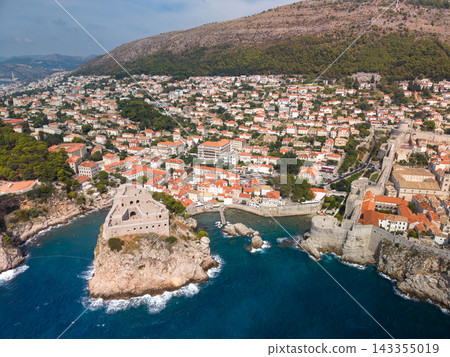 Dubrovnik Croatia fills hillside above Adriatic Sea, its dense terracotta rooftops framed by green slopes. Historic stone walls and coastal fortress define this broad aerial city view. Dubrovnik Croatia fills hillside above Adriatic Sea, its dense terracotta rooftops framed by green slopes. Historic stone walls and coastal fortress define this broad aerial city view. 143355019