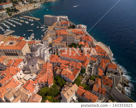 Dubrovnik Croatia fills aerial view, with terracotta rooftops packed inside historic stone walls beside deep blue Adriatic Sea. Old harbor holds clustered boats, framed by coastal fortifications. Dubrovnik Croatia fills aerial view, with terracotta rooftops packed inside historic stone walls beside deep blue Adriatic Sea. Old harbor holds clustered boats, framed by coastal fortifications. 143355025