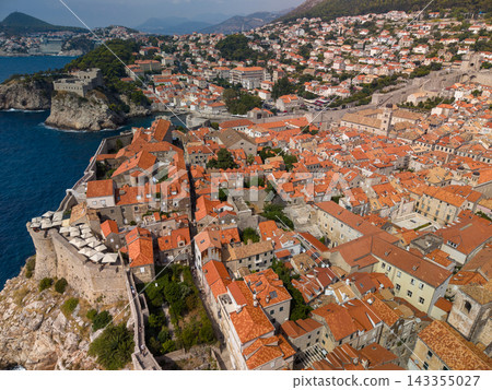 Dubrovnik Croatia spreads across coastal hillside, its old town clustered behind massive stone walls. Orange roof tiles, blue Adriatic water and rocky fortress outcrops shape view. Dubrovnik Croatia spreads across coastal hillside, its old town clustered behind massive stone walls. Orange roof tiles, blue Adriatic water and rocky fortress outcrops shape view. 143355027